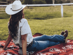 Red and white patterned recycled picnic blanket spread on grass with person wearing white cowboy hat seated on it. preview #2