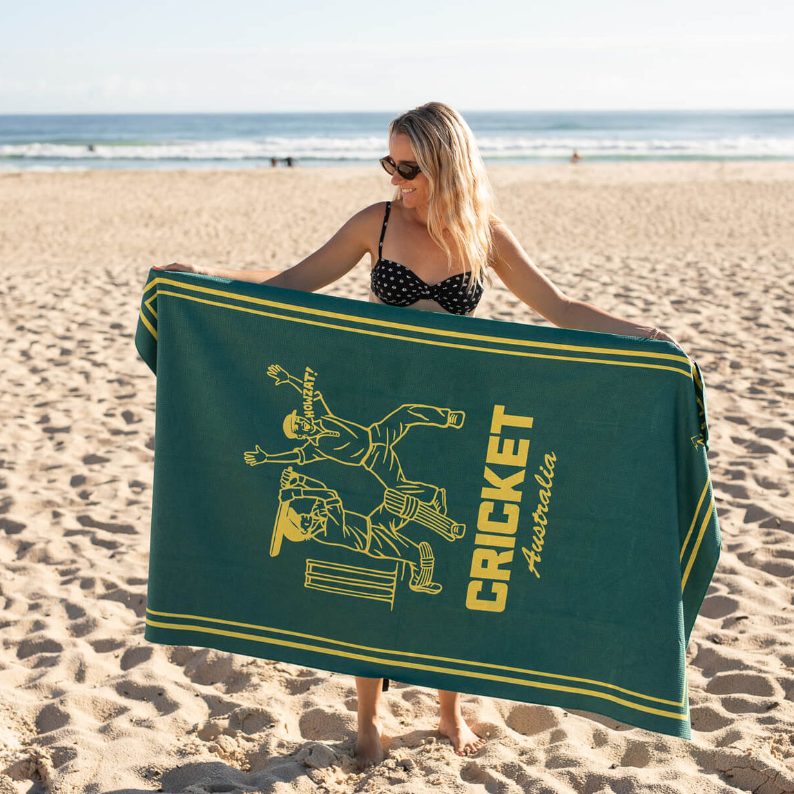 Green and gold beach towel with cricket batter graphic and Cricket Australia text, displayed on sand at the beach