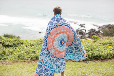 Sustainable down puffy blanket with blue and coral mandala pattern, worn as wrap over shoulders by ocean.
