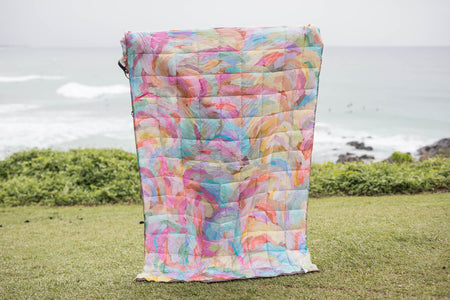 Colorful quilted puffer blanket with watercolor floral print in pink, blue, yellow, and green tones, displayed outdoors.