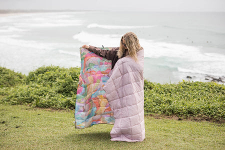 Lavender quilted puffy blanket wrapped around person holding colorful patterned blanket outdoors by ocean