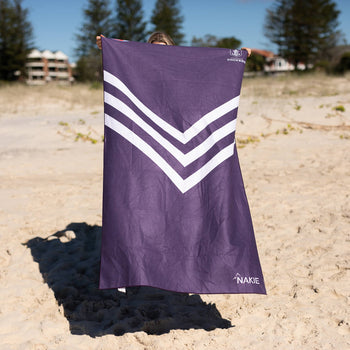 Purple beach towel with white chevron stripe design, held up on sand with beach in background