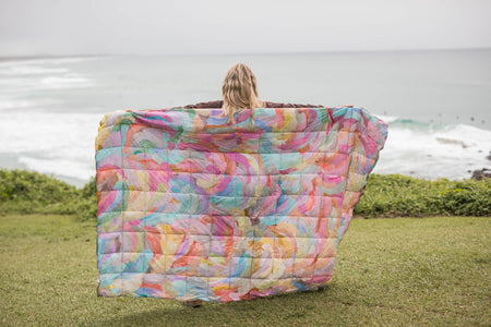 Multicolored quilted puffy blanket with abstract watercolor print, displayed open on grass overlooking ocean.