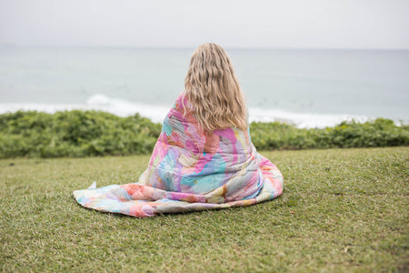 Multicolour tie-dye puffer blanket wrapped around person sitting on grass overlooking ocean