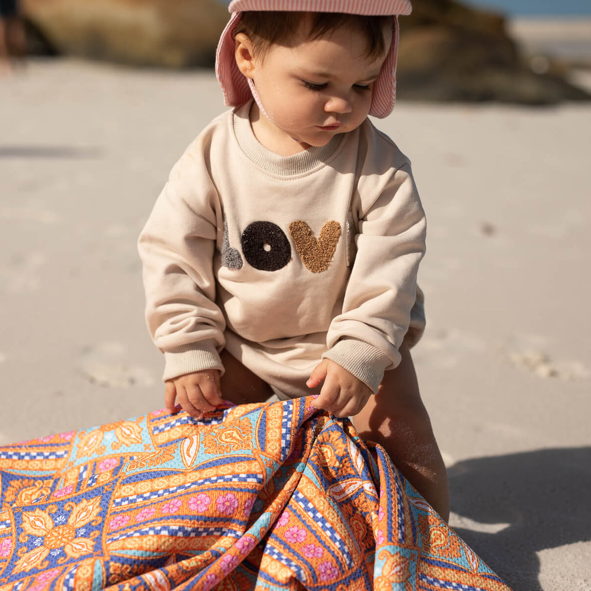 Colorful patterned beach towel in orange, blue, and pink geometric design, held by toddler wearing cream sweatshirt.