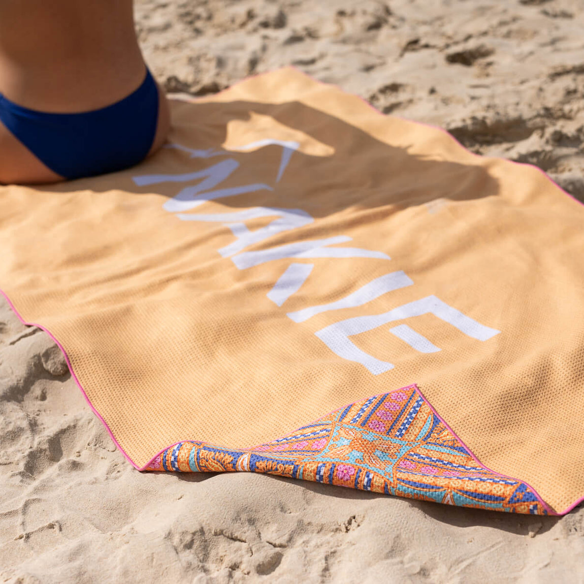 Peach sand-free beach towel with white text and colorful geometric trim, laid on sand with person in blue swimsuit.