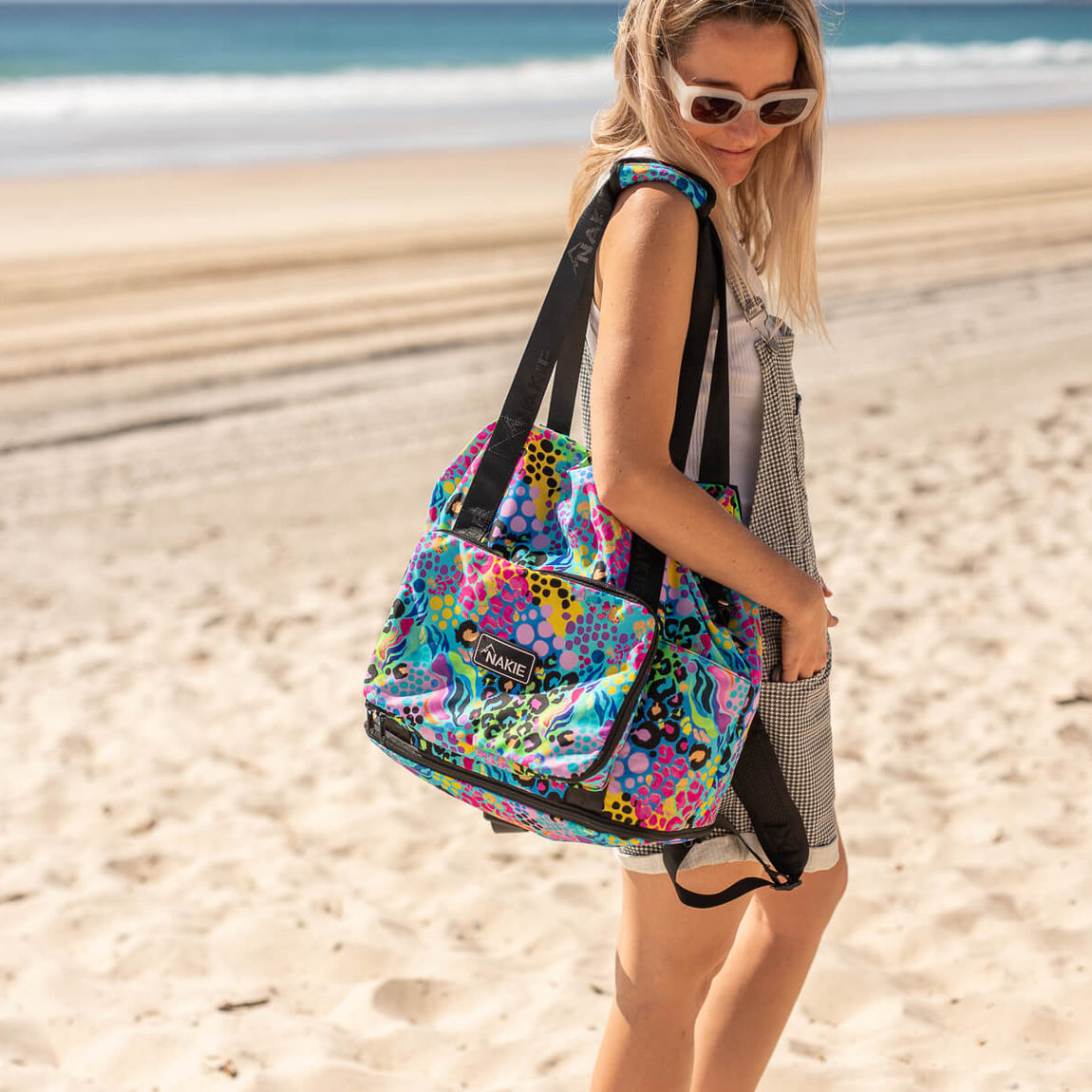 Colorful recycled tote bag with rainbow abstract pattern and black straps, worn on shoulder at beach.