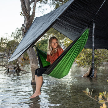 Woman sheltering under a Nakie tarp made from recycled plastic bottles, enjoying a dry outdoor experience.