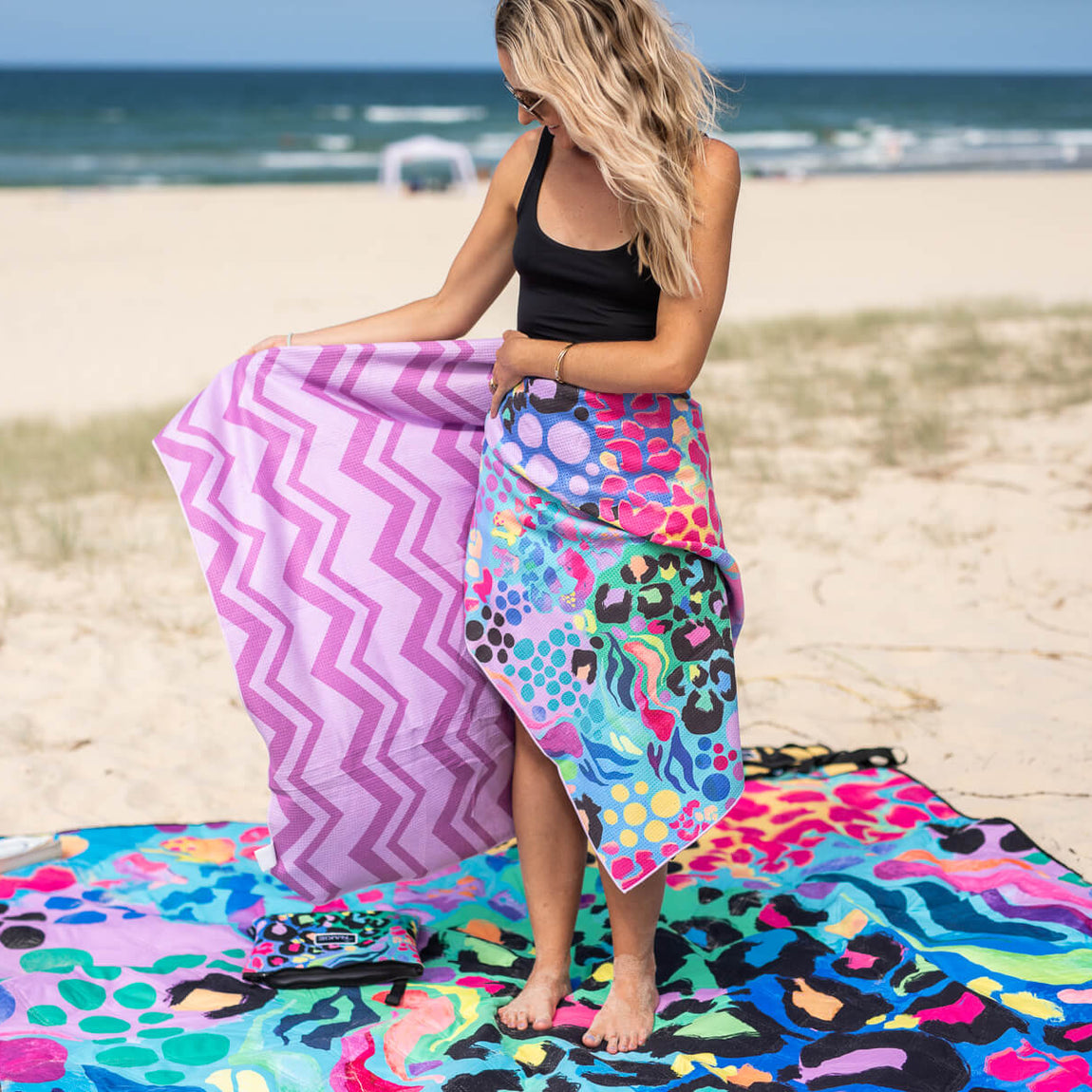 Pink and purple chevron stripe beach towel held by woman standing on colorful patterned sand-free towel at beach.
