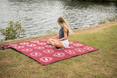 Burgundy picnic blanket with white circular Ringers Western logos spread on grass by water
