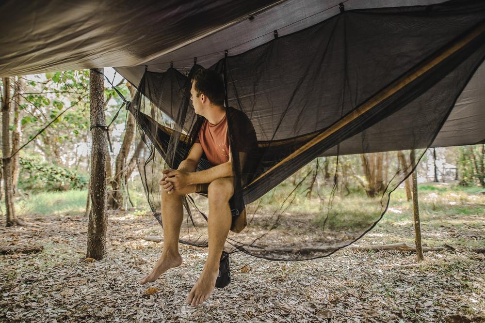 Man lounging comfortably in a Nakie hammock with an integrated bug net and tarp, providing protection from insects and weather elements during outdoor adventures