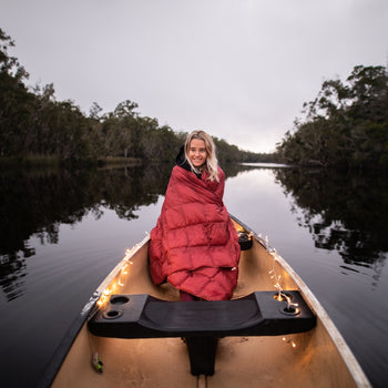 Image of a woman wrapped in Nakie's Earth Red Sustainable Down Puffy Blanket, sitting on a boat and enjoying warmth and sustainability during her maritime adventure.