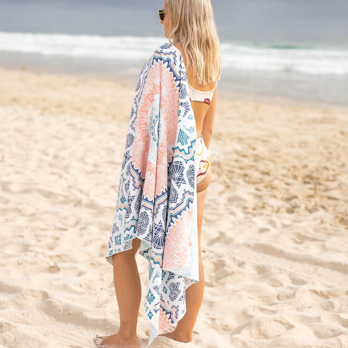 Recycled sand-free beach towel with blue and coral geometric pattern, worn over shoulder on beach