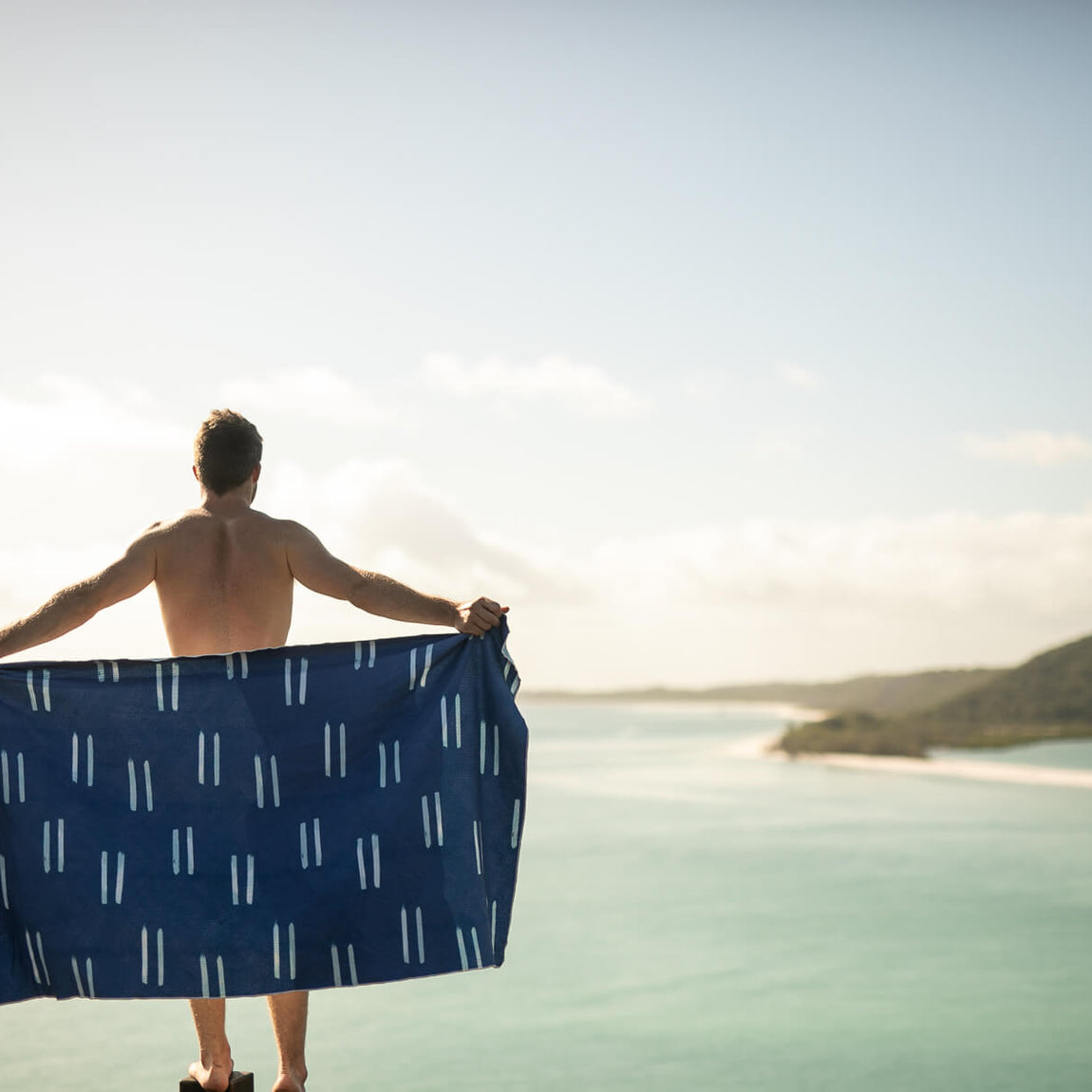 Navy beach towel with white geometric dash pattern, held open by shirtless man overlooking tropical ocean